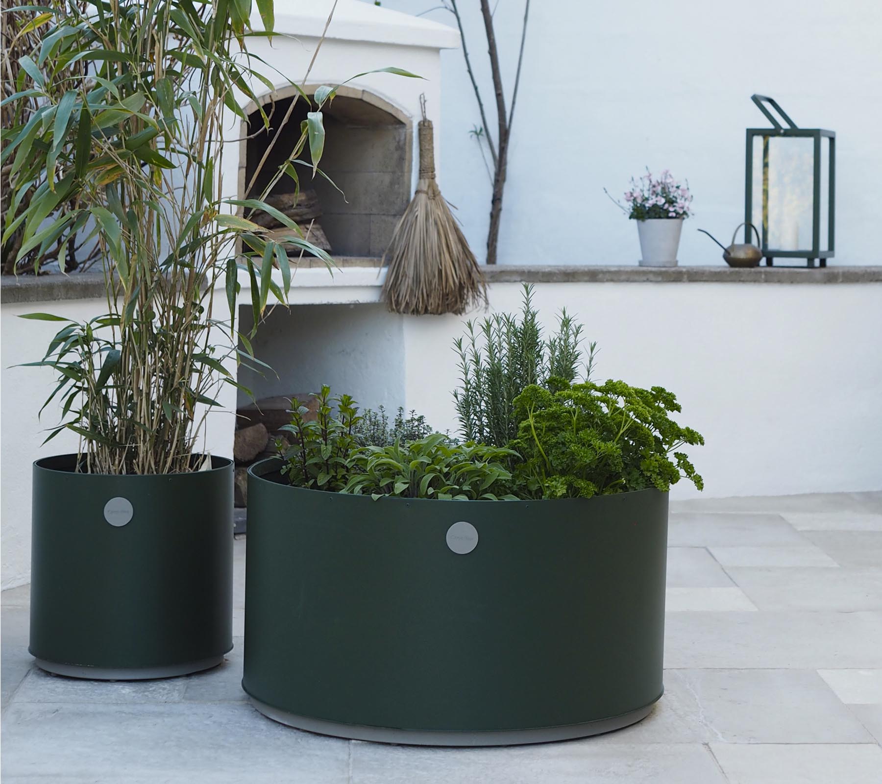 Two round planters in dark green, one featuring tall plants and the other filled with various herbs, sit on a light stone surface.
