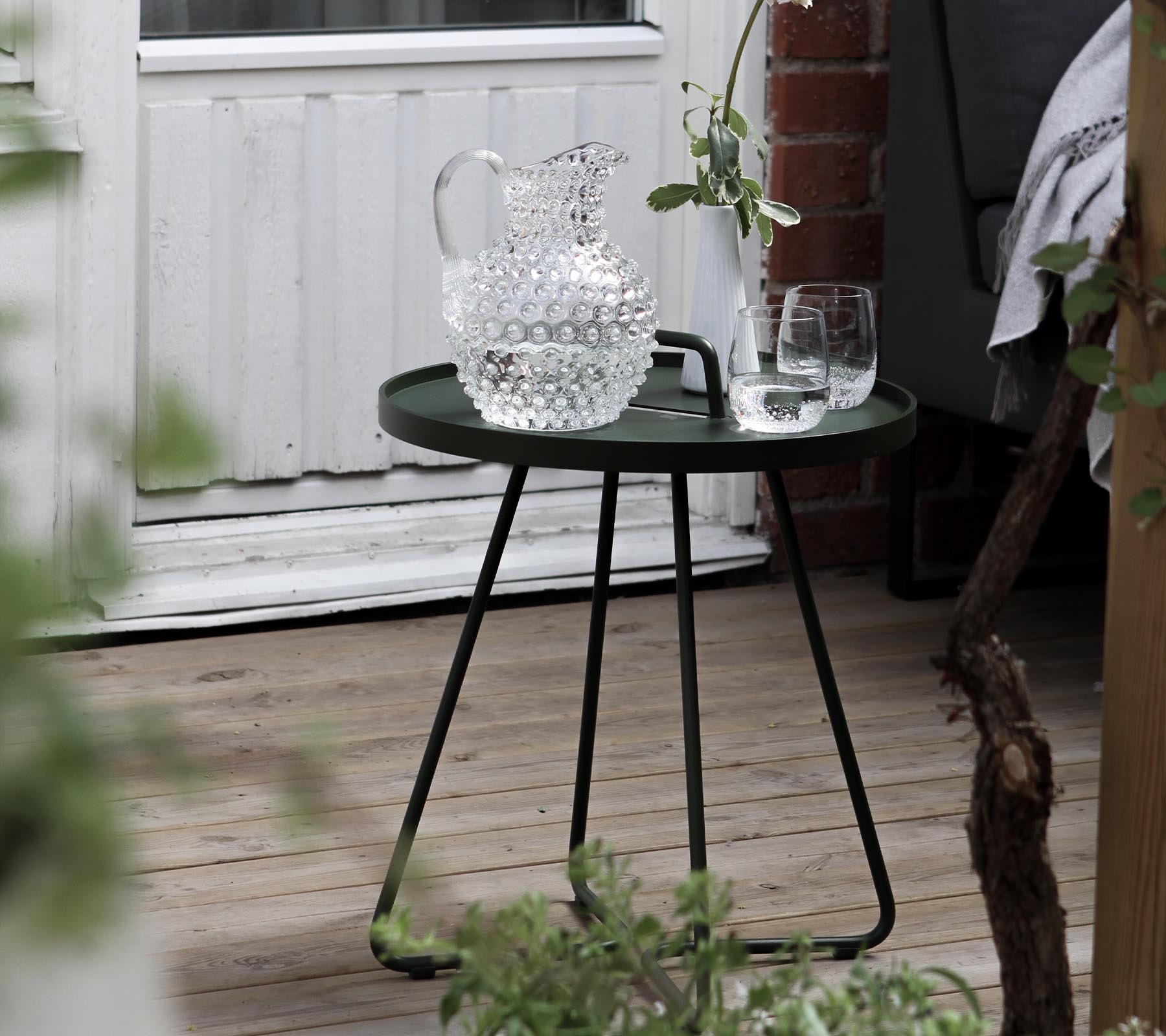 A stylish green side table with a crystal pitcher and two glasses, complemented by fresh greenery in the surrounding area.