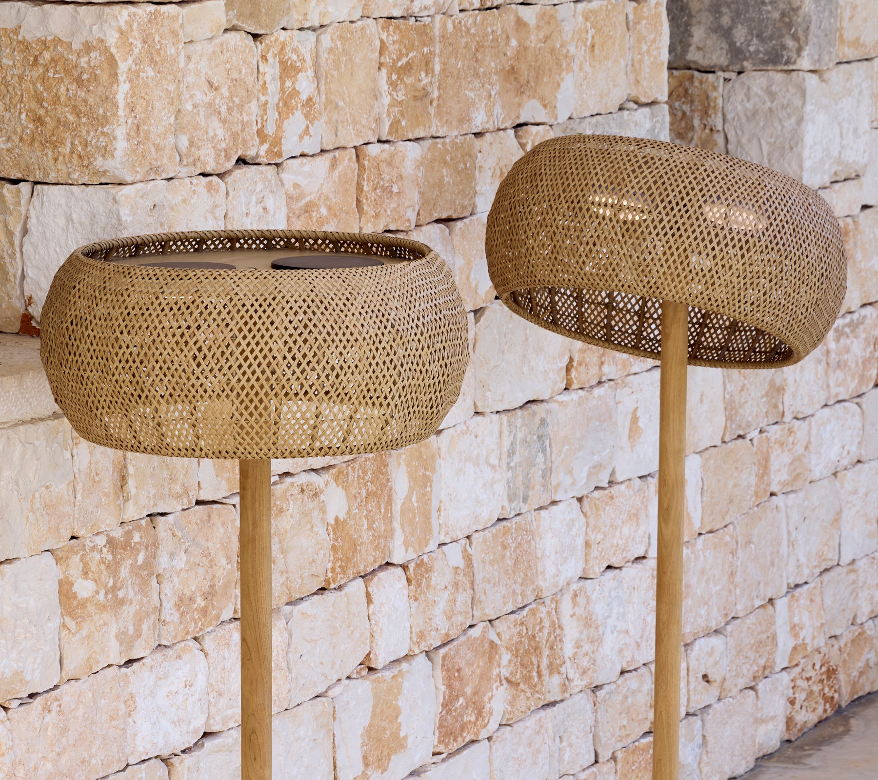 Two woven lampshades on pole lamps against a stone wall.