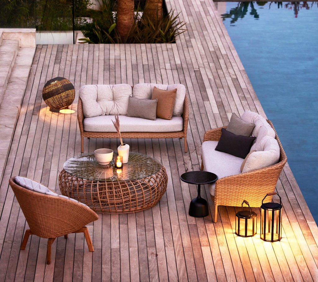 Stylish seating area with woven furniture, a glass table, and decorative lamps, surrounded by wood and reflecting water.
