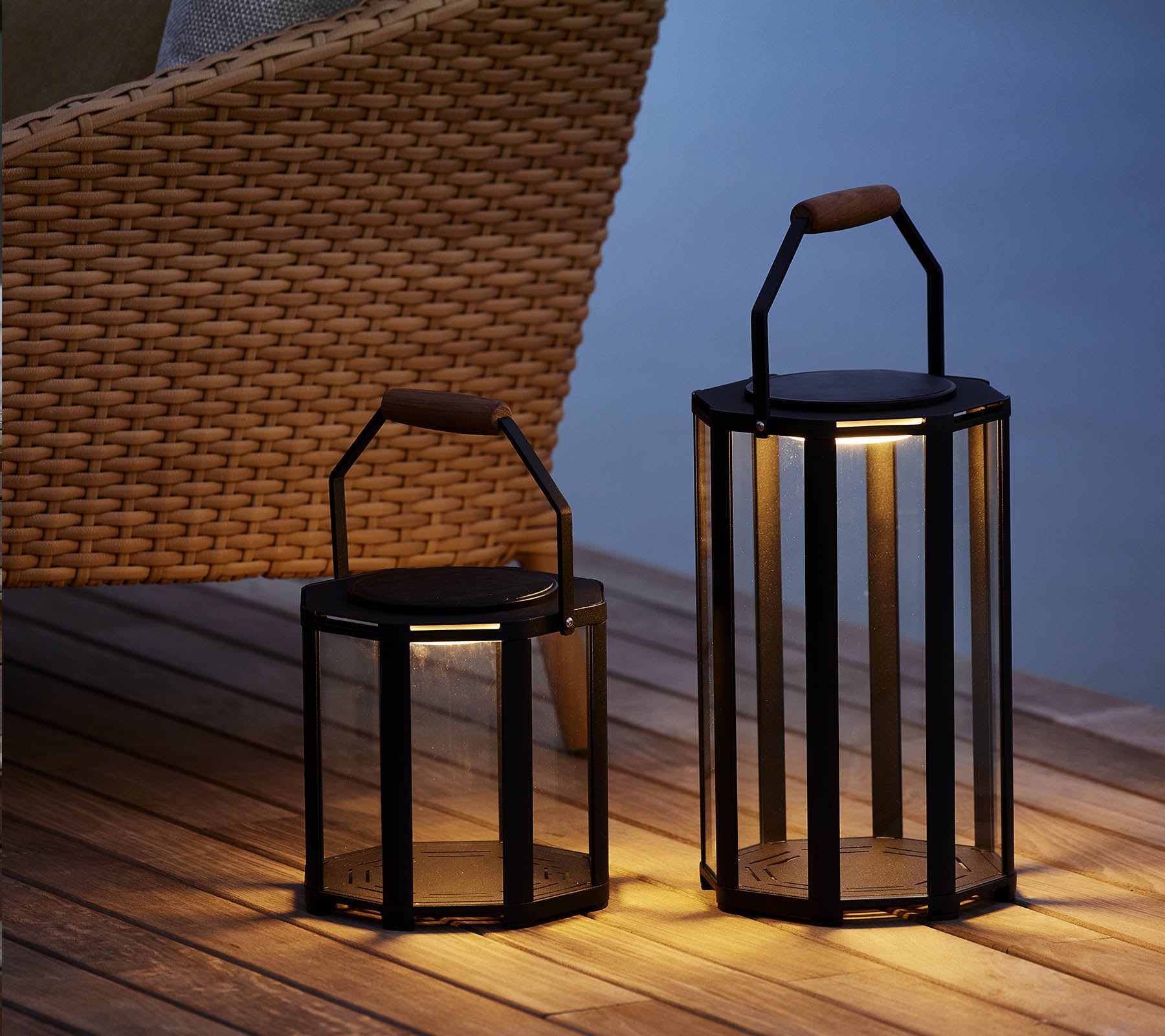 Two stylish Lightluxe lanterns with glass panels and handles, emitting a warm glow, placed beside a woven chair.