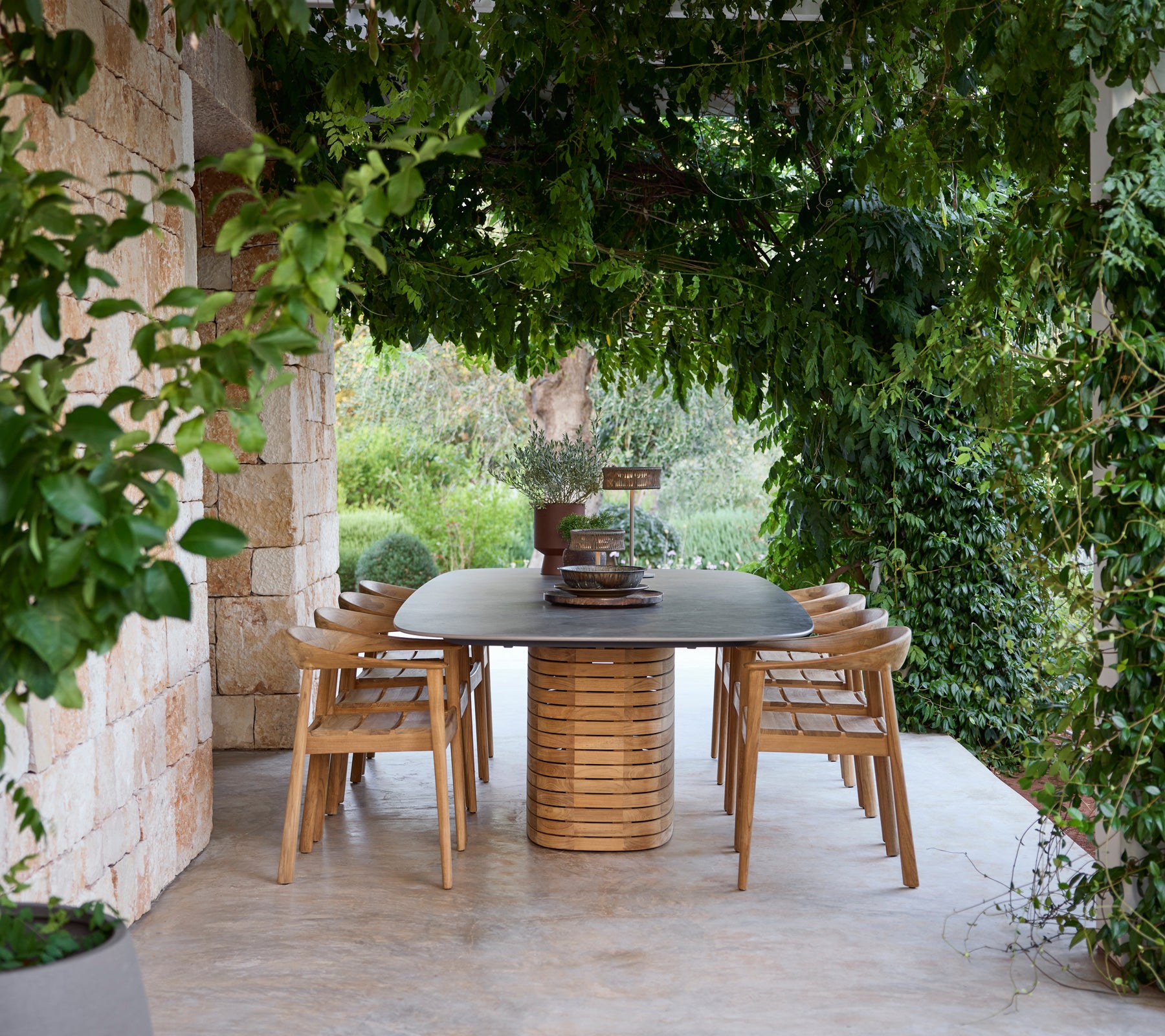 Wooden dining table surrounded by chairs under greenery.