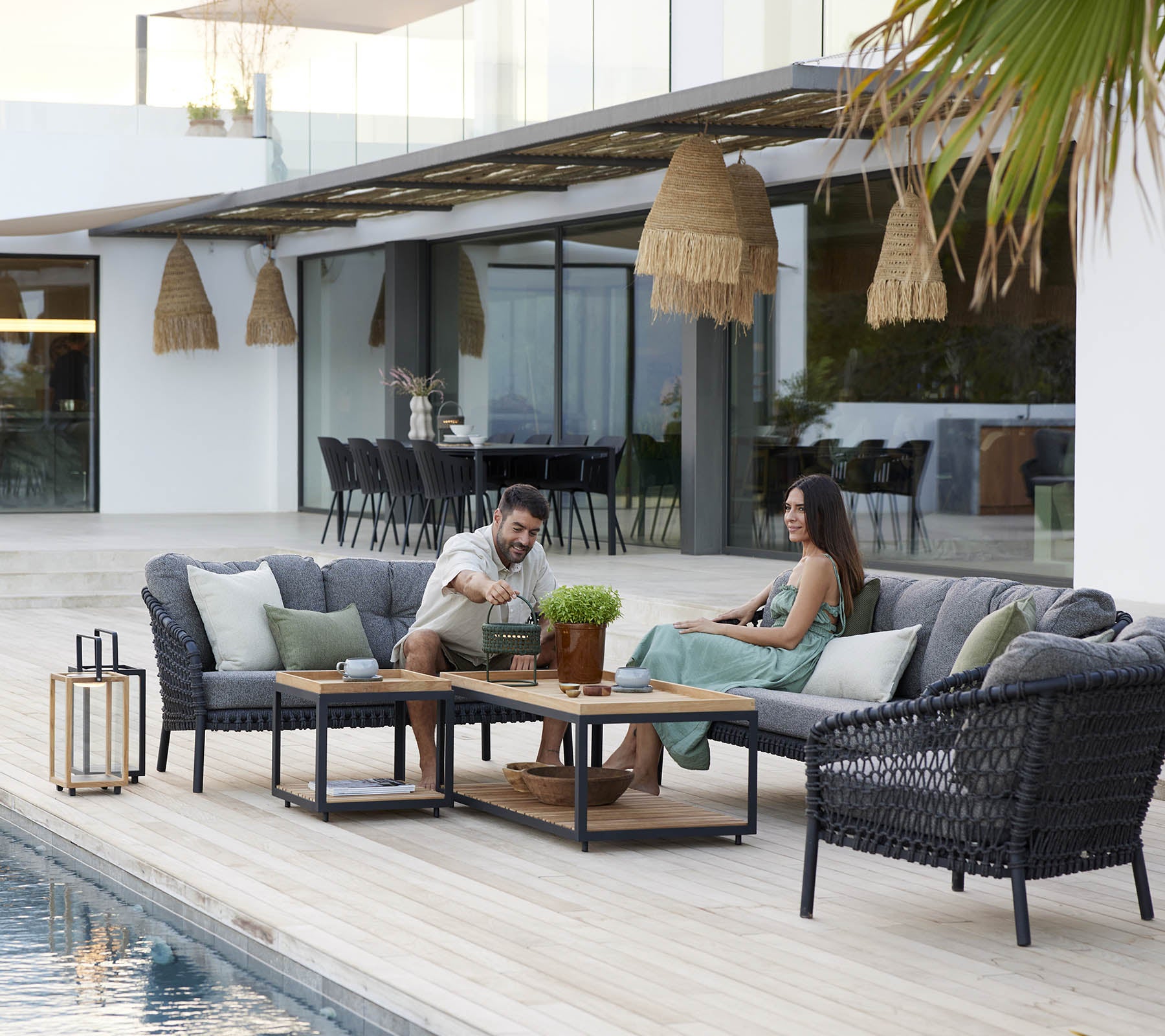 Couple enjoying leisure time on a cozy lounge set, surrounded by green plants and stylish decor, with a serene swimming pool nearby.