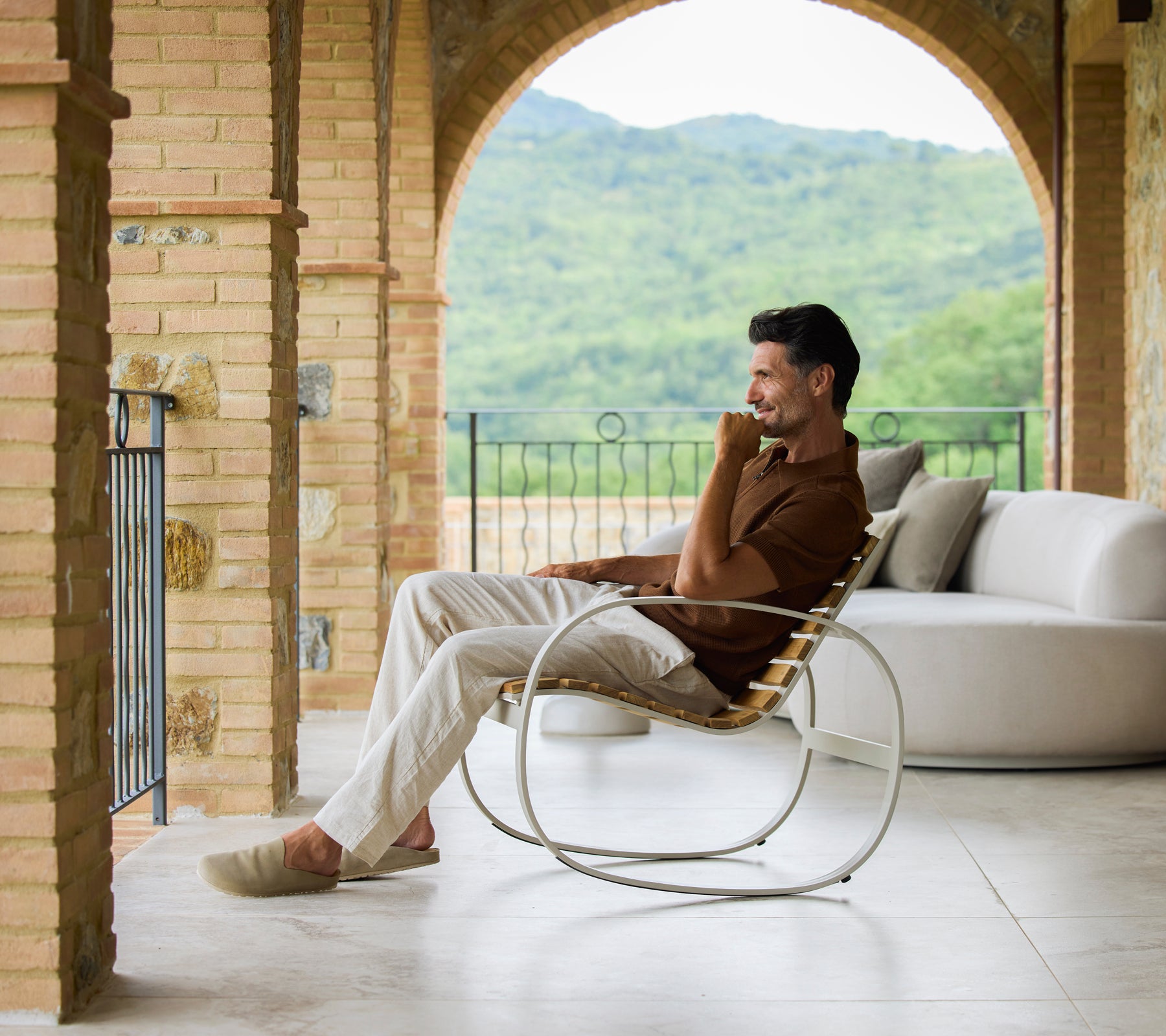 Man relaxing in a sand-colored rocking chair, enjoying scenic views and a serene atmosphere. Soft, neutral tones highlight comfort.