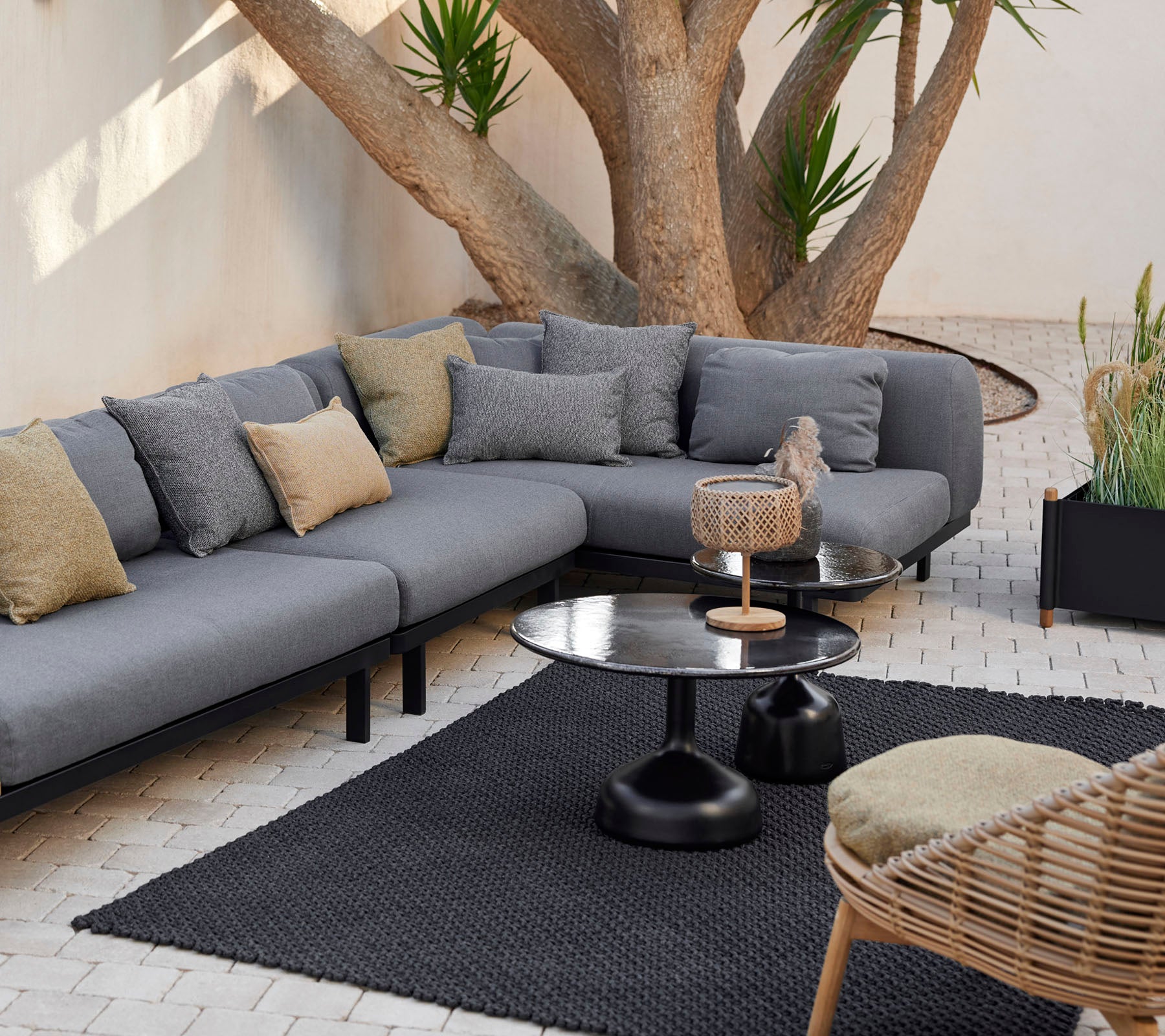 Lounge area featuring a grey modular sofa with decorative pillows, a sleek coffee table, and a textured rug surrounded by greenery.