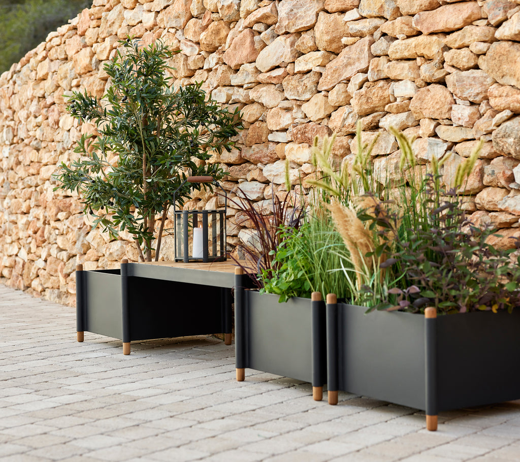 A stylish bench with two planters filled with greenery, accompanied by a lantern, set against a stone wall.
