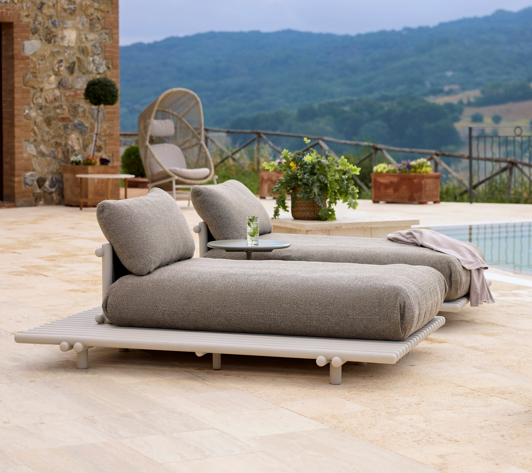 Stylish aluminium sunbeds with soft cushions, accompanied by a small table, set against a scenic backdrop of greenery and hills.