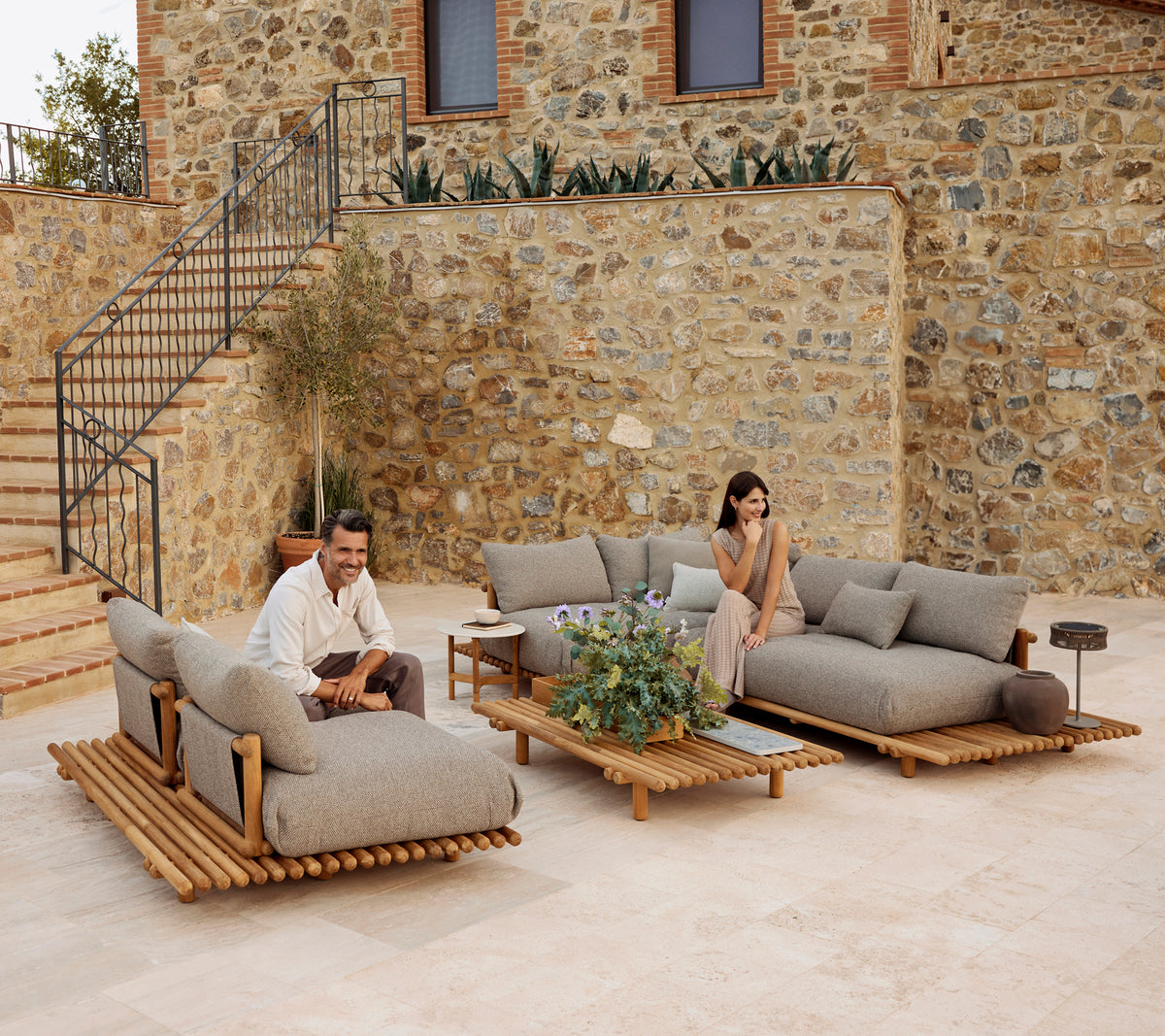 A stylish lounge area featuring teak furniture with plush gray cushions, accented by greenery and a modern coffee table.