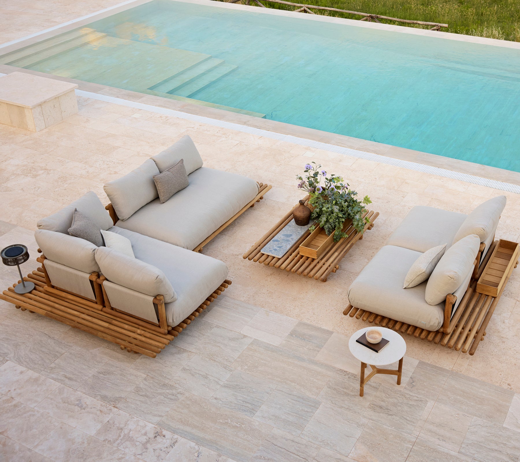Modern outdoor seating area featuring neutral cushions, wooden platform tables, and a small potted plant beside a pool.