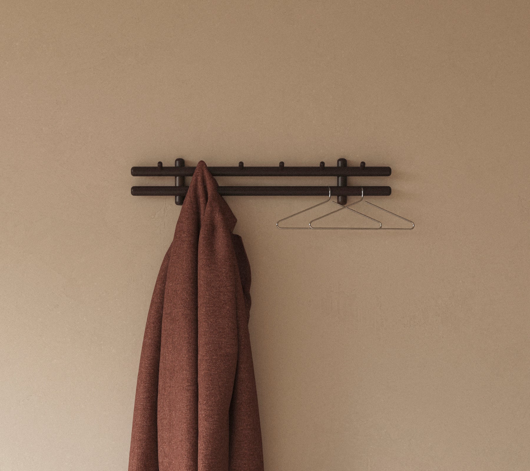 A brown coat hangs from a minimalist wall rack with hooks, complemented by a simple wire hanger against a neutral background.