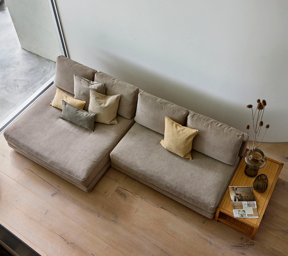 A stylish sofa in a neutral tone, adorned with various decorative pillows, accompanied by a wooden side table with books and a vase.