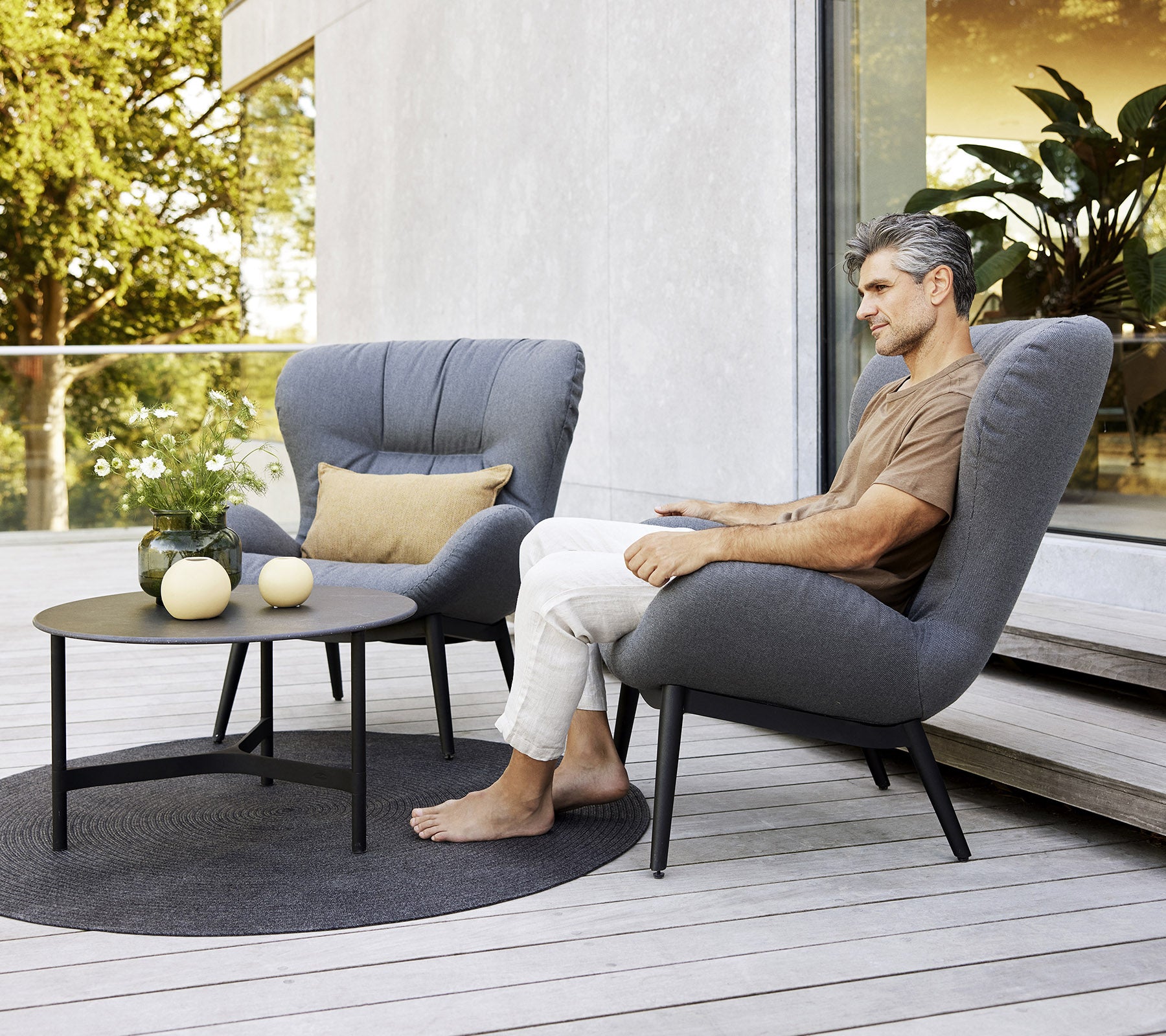 A man sits in a cozy chair next to a small table with decorative items, surrounded by greenery in a serene setting.